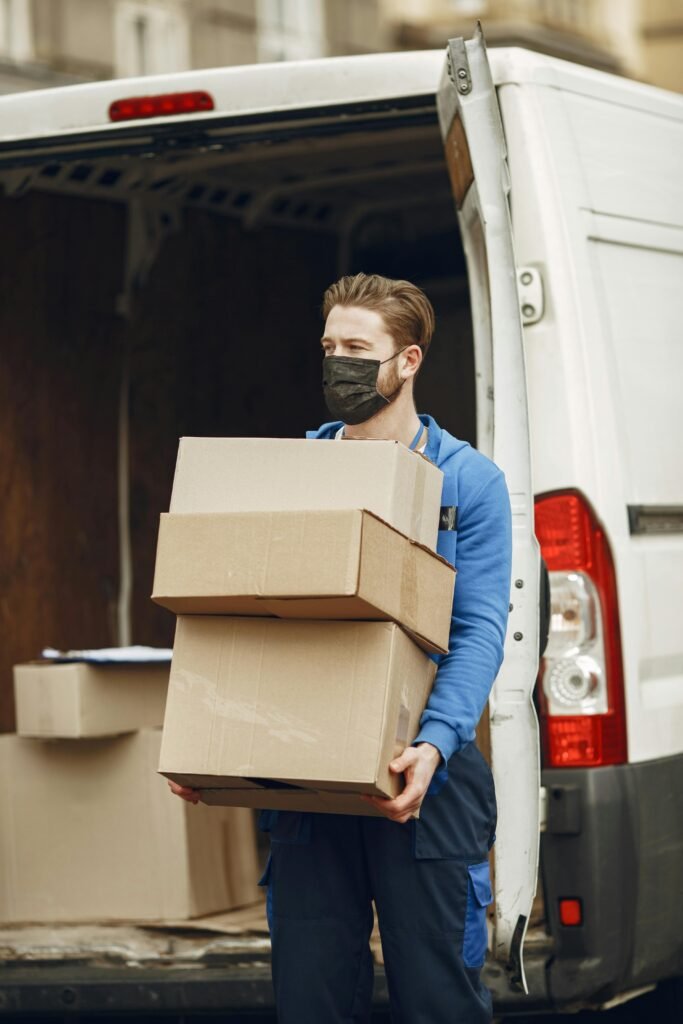 Man wearing mask delivering cardboard boxes from van outdoors.